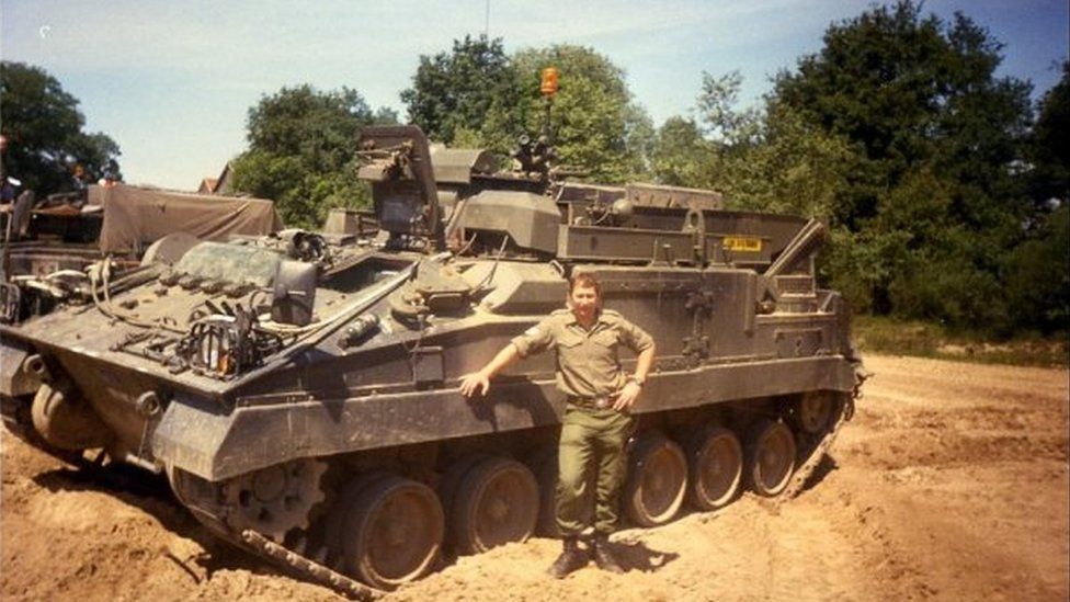 Peter Kay standing in front of an army tank