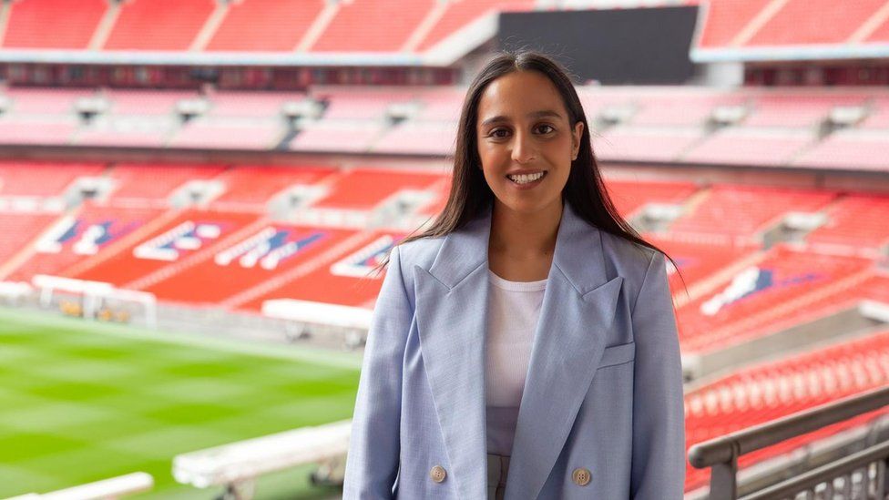 Hina Shafi wearing a light blue blazer and stood smiling inside Wembley Stadium which is behind her