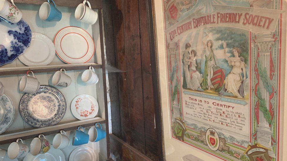 fancy dishes on a shelf beside a Victorian poster fro m the Equitable Friendly society