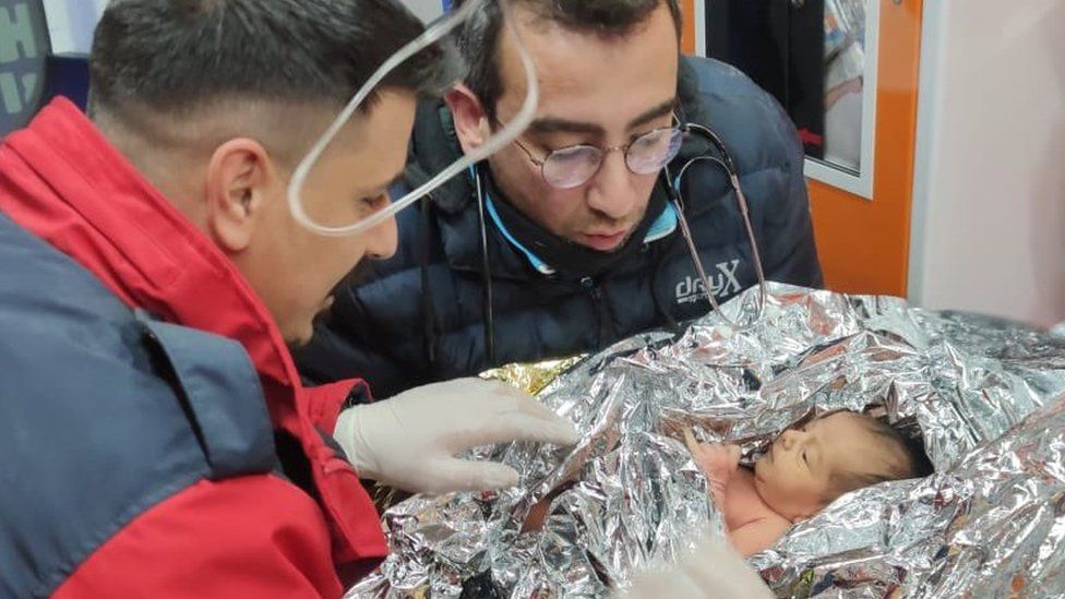 Un bebé envuelto en una manta térmica es atendido por dos hombres tras ser rescatado de un edificio en ruinas