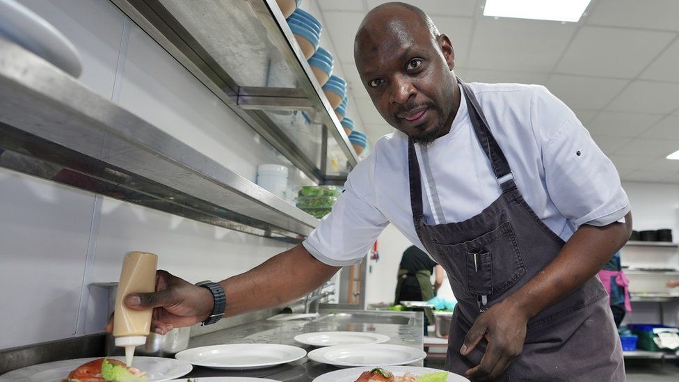 Chef George Opondo looks at the camera as he applies garnishes to dinner plates in a large kitchen