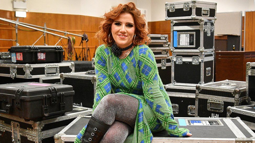 Ciara Mary-Alice Thompson, known as CMAT poses at BBC Radio 1's Sound of 2024 LIVE at BBC Maida Vale Studios in January 2024. CMAT is a 27-year-old white woman with shoulder length curly red hair. She wears a green and blue checked dress over sparkly grey tights and knee-high black boots. She poses sat on a black and silver equipment box and is surrounded by multiple huge boxes in the studio.