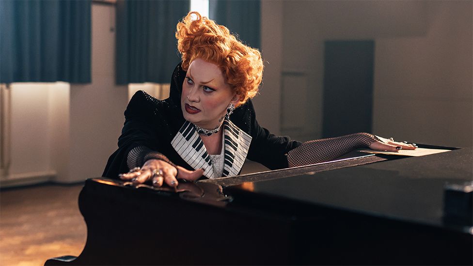 A character in an elaborate costume sits at a piano, looking at someone or something out of shot with something like disdain. They have a hand placed on top of the instrument at each end, giving us a glimpse of their elbow-length fishnet gloves. They wear a black jacket with an oversized collar printed with a piano key pattern. They wear a large silver chain around their neck, and their red hair is styled in an Elizabethan perm.