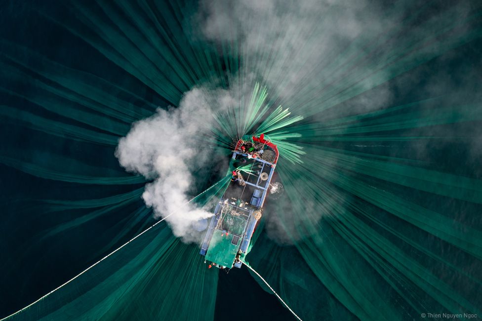 An aerial view of smoke rising from a boat, with green nets in the water below