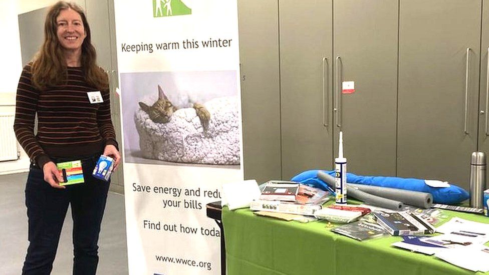 Image of Jessica Thimbleby at an Energy Cafe in Malmesbury. She is pictured standing next to a table full of leaflets and items such as blankets and light bulbs