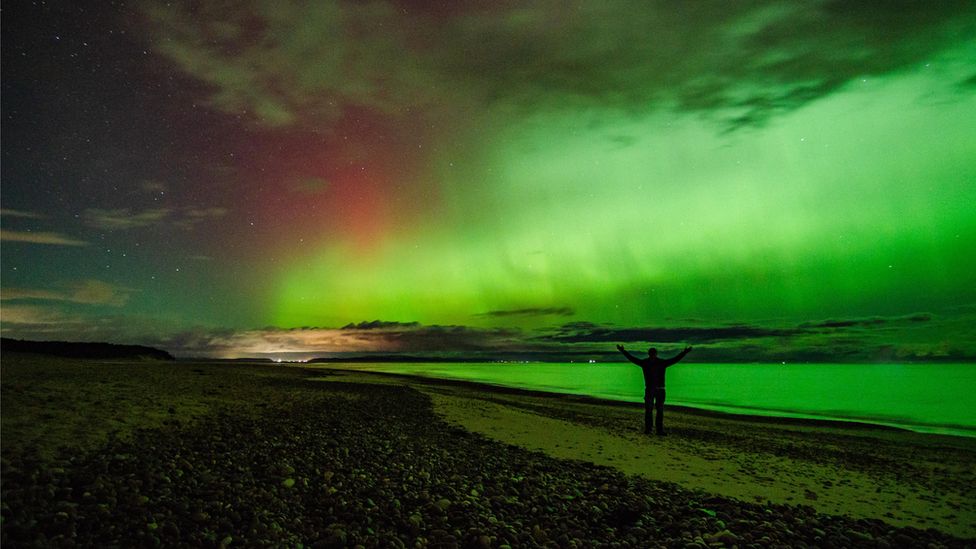 The Northern Lights seen from a beach with a person with arms outstretched standing on the beach