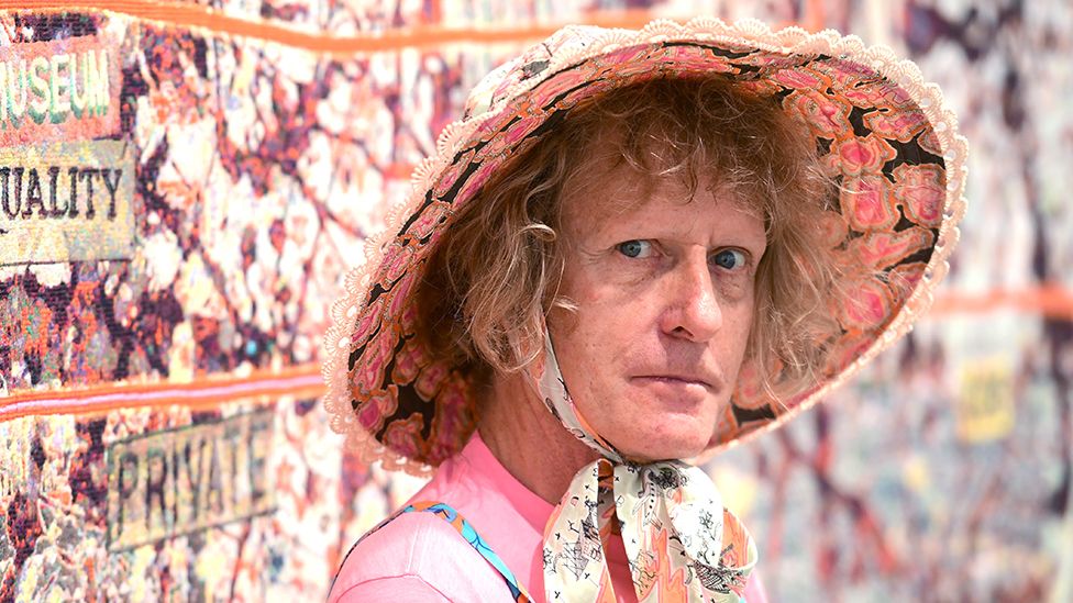 Grayson Perry looking at the camera wearing a hat in front of an artwork, taken in 2020