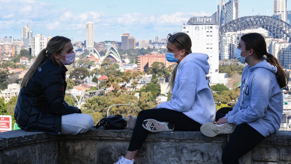 Three girls wearing face masks sit above the Bradfield Highway in the suburb of North Sydney with the city in the background on July 07, 2021 in Sydney, Australia.