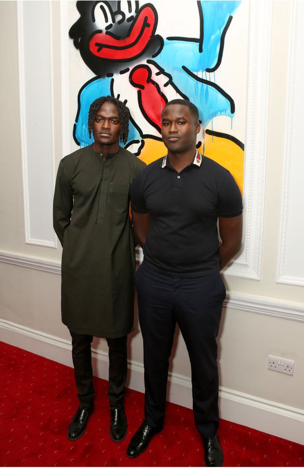Two men wearing black standing in front of a colourful piece of work, which appears to show a figure in black face, with large red lips.