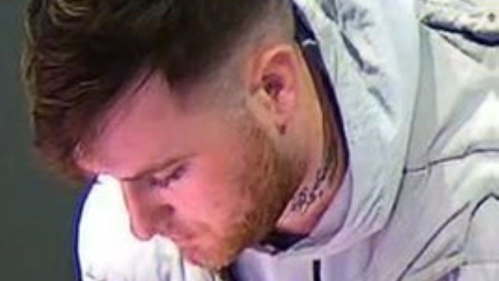CCTV image of a young man with brown hair, facial hair with a tattoo on his neck. He is wearing a white or grey hooded top or jacket.