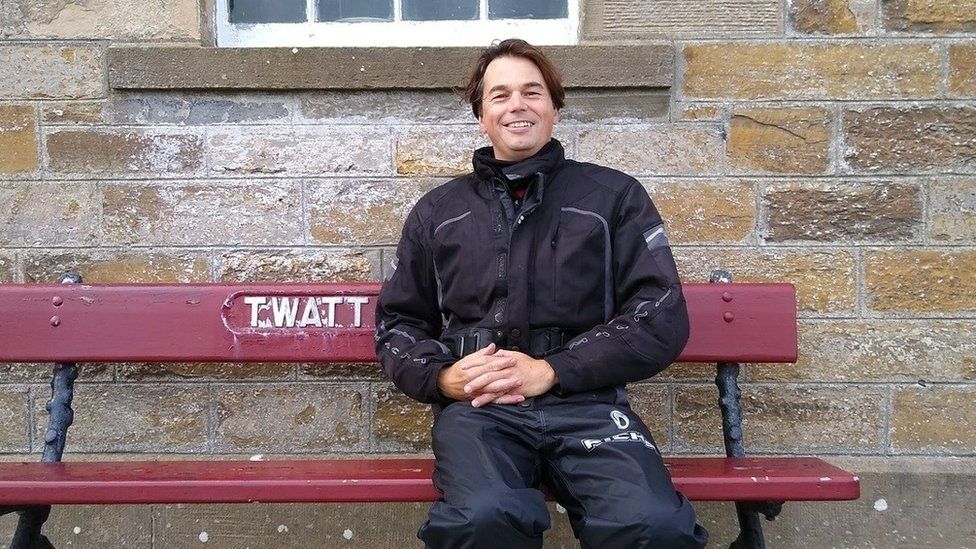 Paul Taylor sitting on a bench with the place name of Twatt painted on it