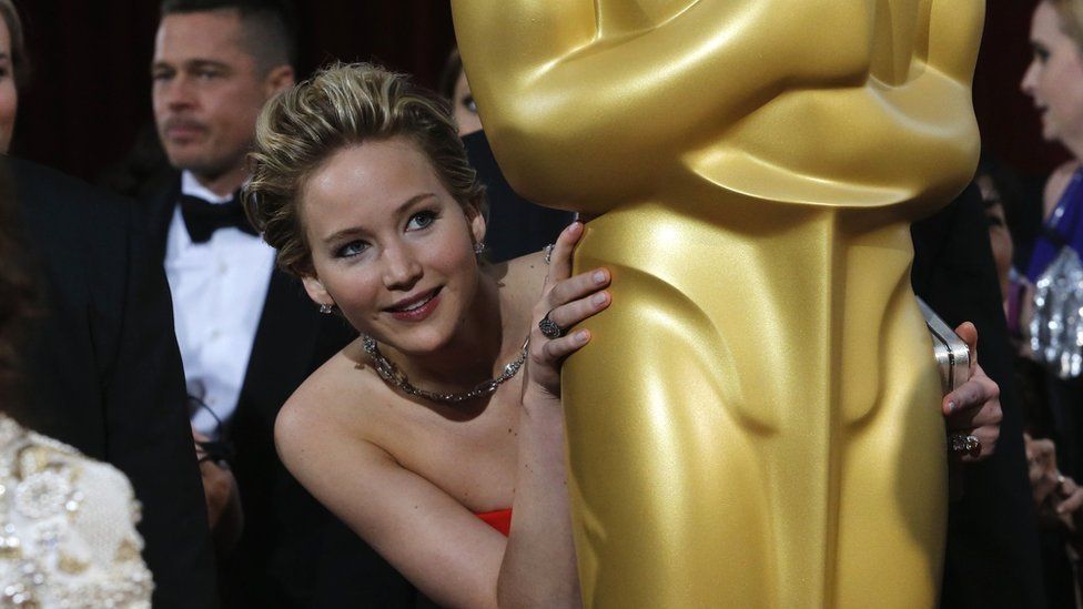 Jennifer Lawrence hiding behind an Oscar award statue.