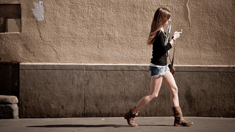 Уличная фотография, женщина гуляет, уделяя внимание мобильному телефону.