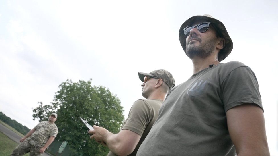 Anton, um operador de drone, está do lado de fora com chapéu e óculos escuros, com um homem operando um drone atrás dele
