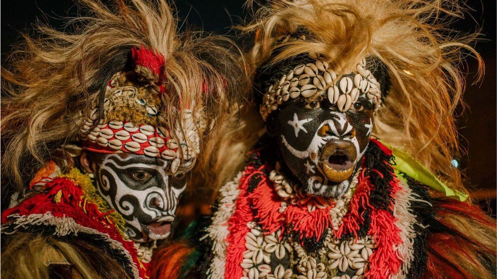 Two men with white face paint on, wearing elaborate lion-like head dresses and colourful accessories.