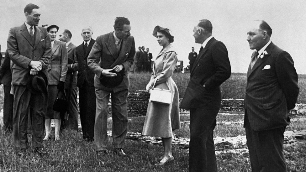 Black and white image of the Queen in 1952 surrounded by men in suits