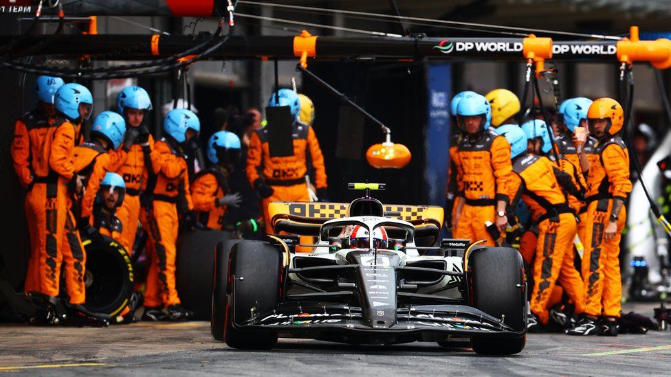 McLaren F1's Lando Norris in his car leaving a pit stop, with his team behind him watching on, dressed in orange racing suits with a mix of orange and blue helmets.