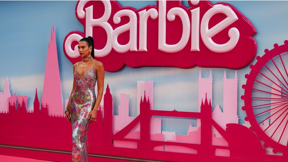 Поп-звезда Дуа Липа позирует на розовой дорожке премьеры фильма о Барби. На ней разноцветное блестящее платье с ремешками, а волосы собраны в тугой хвост. Она стоит перед Барби на фоне горизонта Лондона.