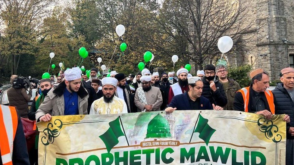 Marchers hold a banner stating "Prophetic Mawlid" and hold blue and green balloons.