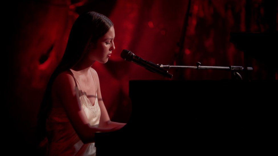 Olivia Rodrigo in the Live Lounge. Olivia is a 20-year-old woman with long brown hair worn loose over her shoulders. She wears a white silk slip-style dress with a lace trim and has red lipstick on. She sits at a piano, her hands on the keys, and sings into a microphone. Behind her the backdrop is dark and coloured red.