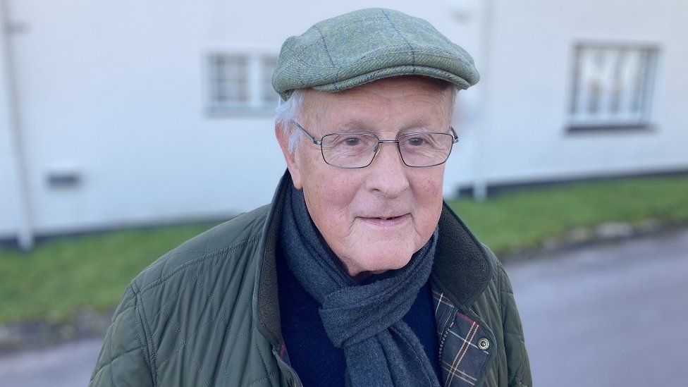 Derek Finnis looks at the camera, wearing glasses, a flat cap and winter coat