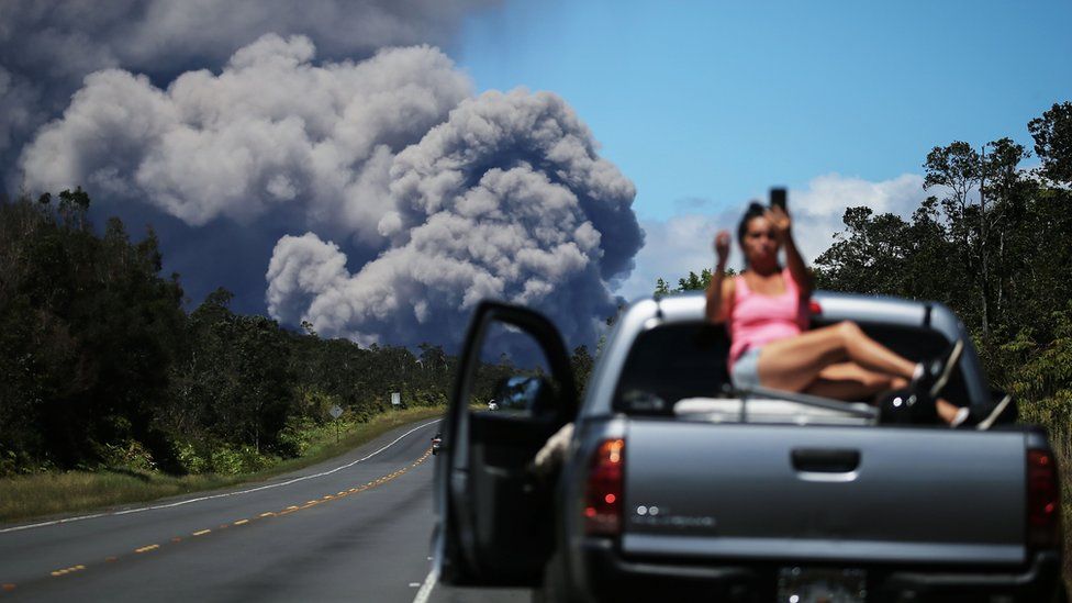 A woman takes a selfie with a cloud from Kilauea in the background