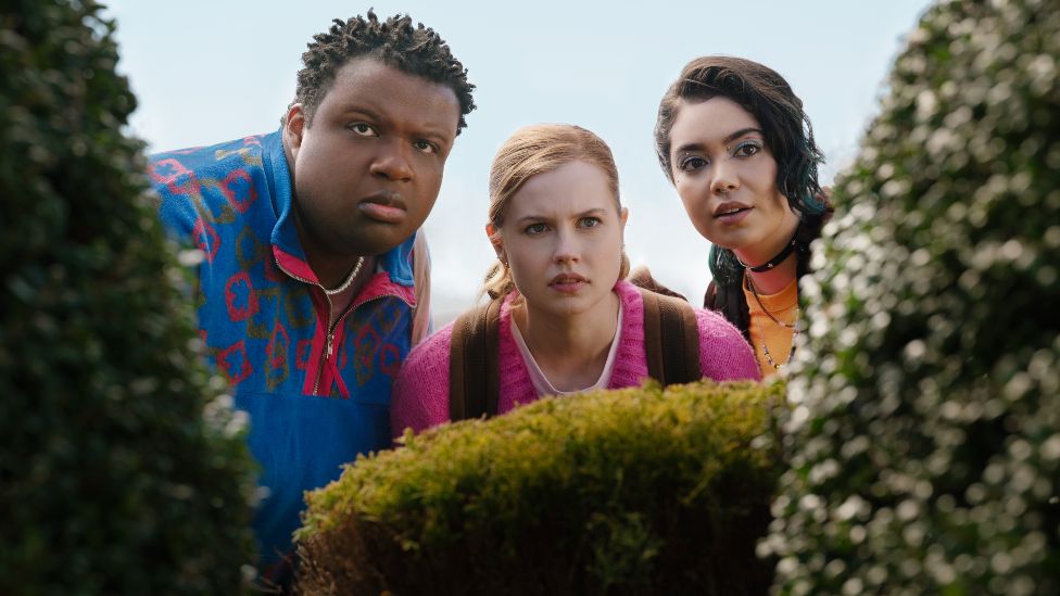 Promotional still from 2024 film Mean Girls. L-R is Jaquel Spivey as Damian, Angourie Rice as Cady and Auli'i Cravalho as Janis. The trio are pictured hiding behind a hedge - Damian, the tallest of the three, wears a bright blue patterned fleece, Cady wears a brown backpack over a pink jumper and Janis wears a high-necked orange top