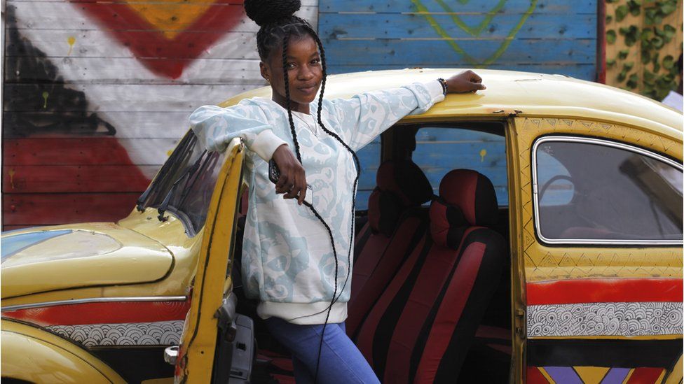 A young woman standing next to an old yellow Volkswagen. There are red, black and white stripes on the car.
