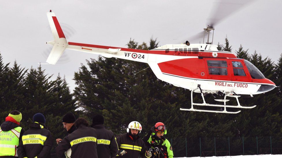 A red rescue helicopter is seen landing next to workers in high-vis snow gear