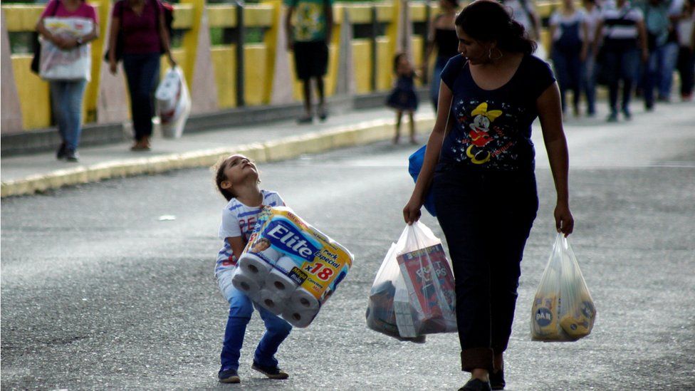 a child struggles with an 18-pack of toilet paper next to a woman carrying multiple plastic bags, 10 July 2016