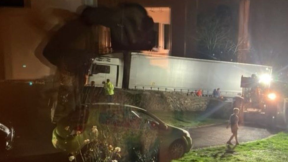 A slightly blurred picture in darkness showing a big white lorry behind a wall on a tight road, with people around trying to help them out.