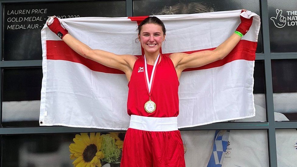 Elise Glynn, a white, brunette woman, wears a red boxing uniform and holds an England flag above her head. She is smiling and wearing a medal around her neck