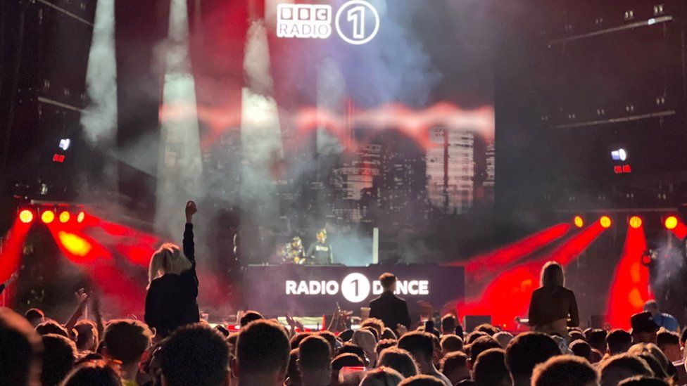 A crowd of people facing the Radio 1 Dance stage at the Big Weekend festival. One woman is on someone's shoulders and has her arm in the air