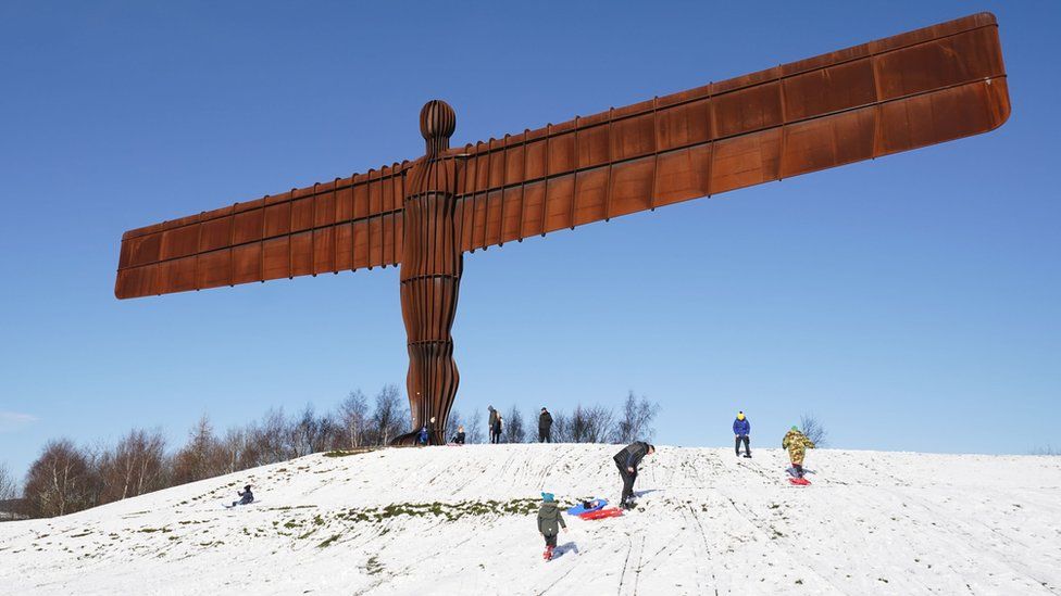 People sledge next to the Angel of the North, a large metal form of a human with enormous outspread arms