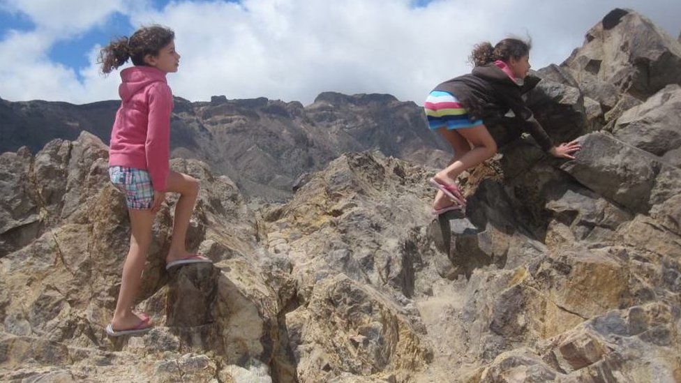 Twin girls clamber across rocks on a mountain with their back to the camera