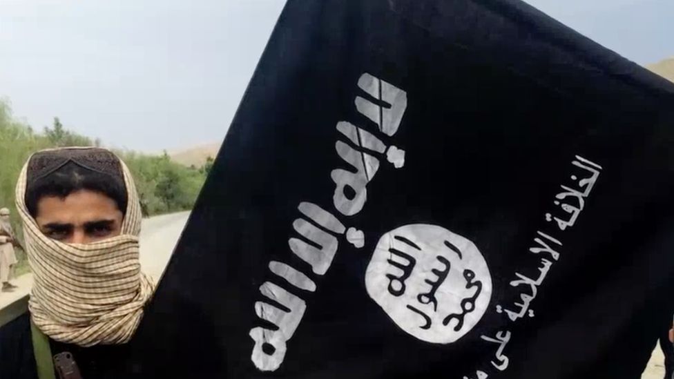 Screenshot of IS video showing a man in a scarf standing next to an IS flag