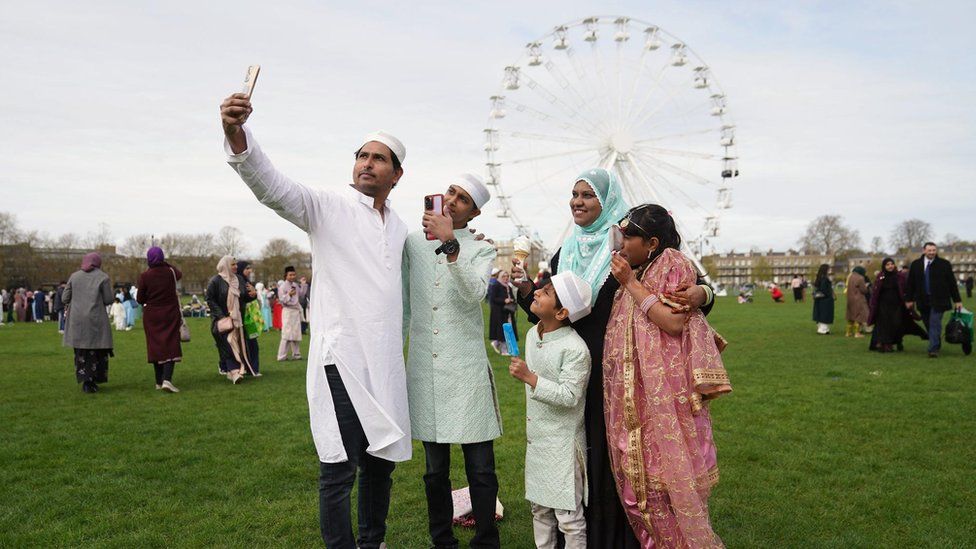 A family take a selfie with an observation wheel in the background, Cambridge, UK.
