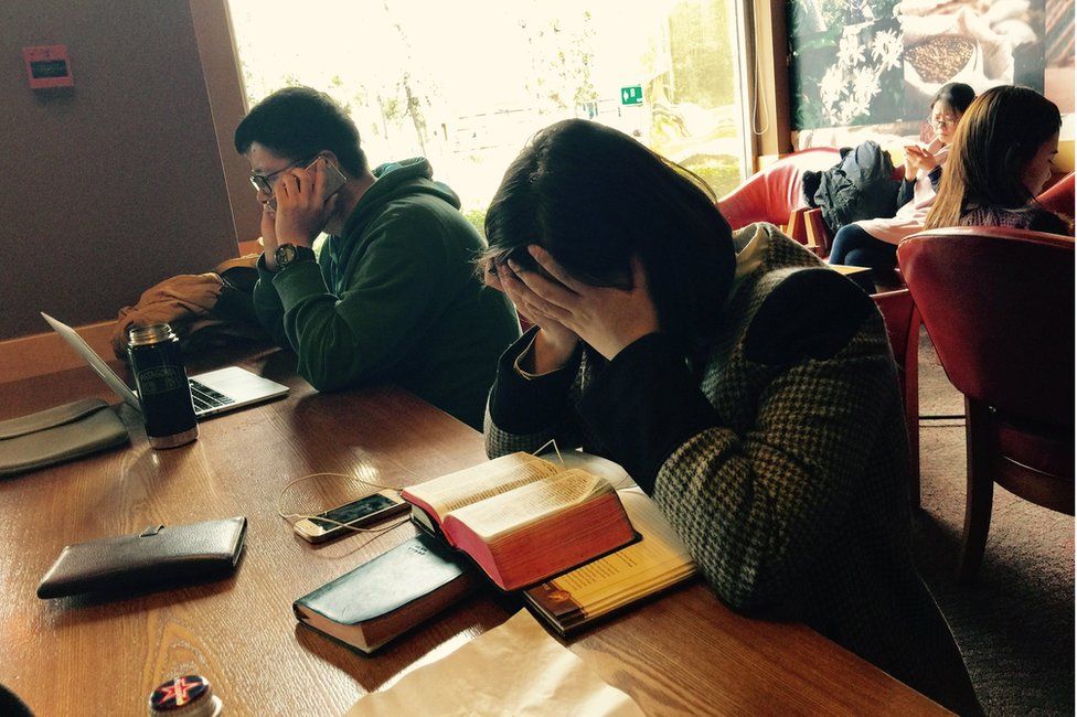 Cafe in China with a woman holding her head in her hands in front of a Bible, and a man on his mobile phone in front of his laptop.