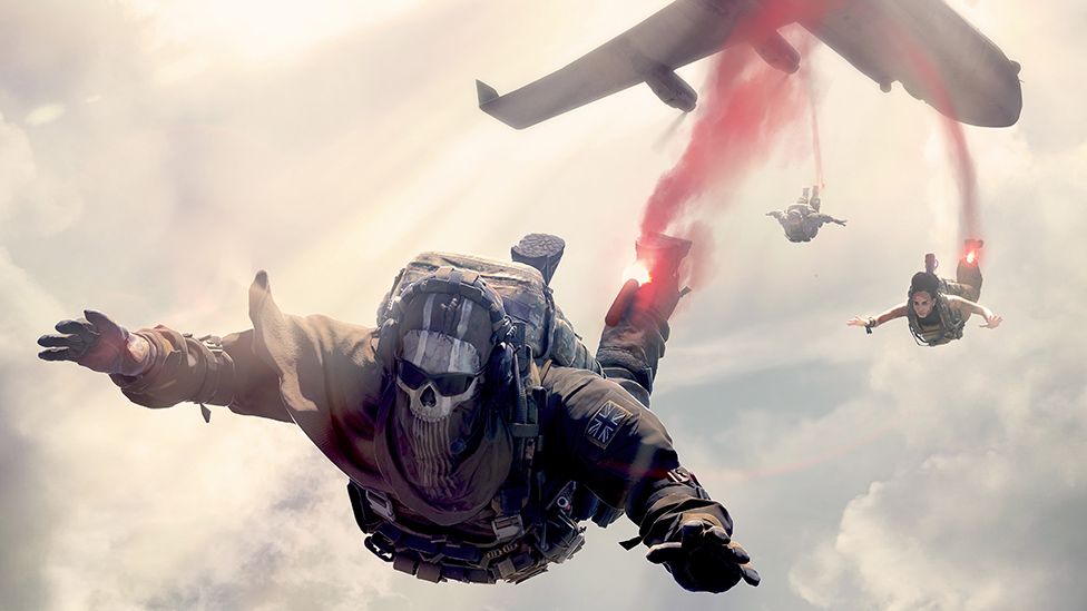 Three figures leap out of a cargo plane into a cloudy sky. They each have a flare attached to their ankle, emitting red smoke which forms a long trail marking the path of their descent. The figure nearest the camera has his arms stretched towards the viewer. he's wearing military clothes, ready for combat, and wears a black face mask with a white skull pattern. Behind him, further away, we can make out a female character and, even further back, a male character.