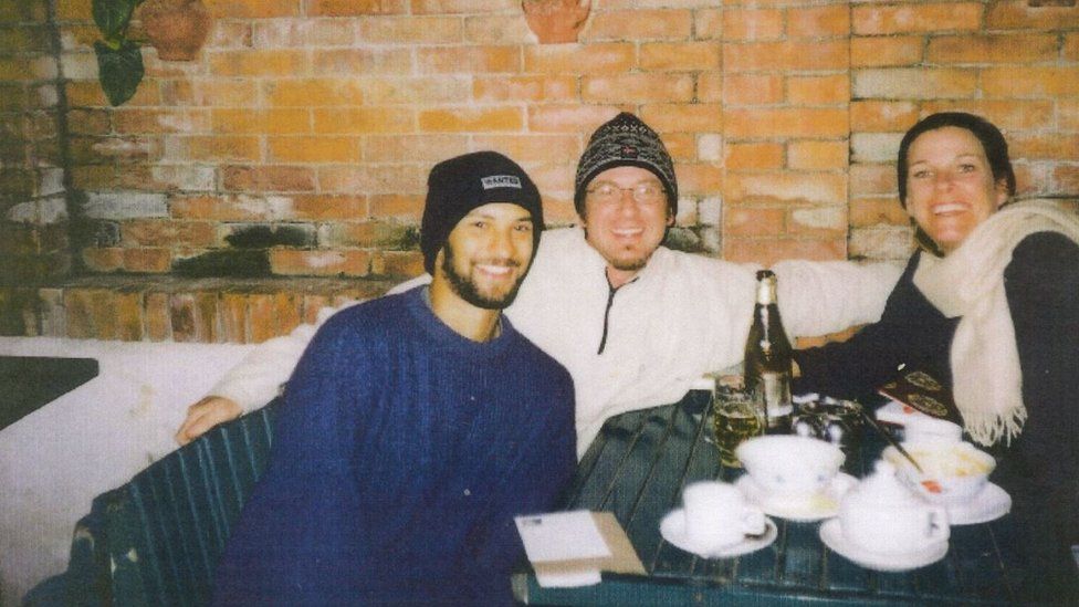 Letztes Foto von Alex in Nepal, er trägt einen schwarzen Hut, einen blauen Pullover und einen Bart. Er sitzt mit zwei anderen Personen an einem Tisch