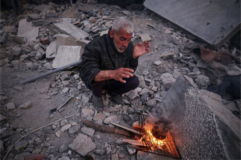 Khaled Naji cooks over a fire in the ruins of his home in Deir al-Balah, Gaza. He is struggling to feed family. (Majdi Fathi/BBC)