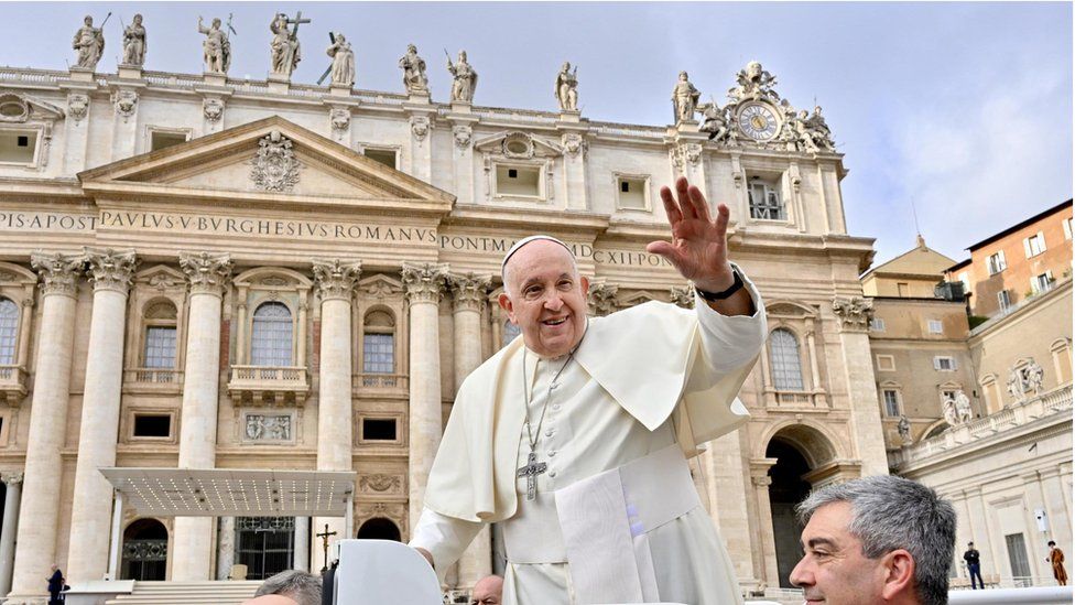 Pope Francis with hand up in front of Vatican building