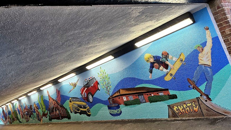 An underpass with a mural on one side, with a background in different shades of blue and skateboarders, cars and dragons painted