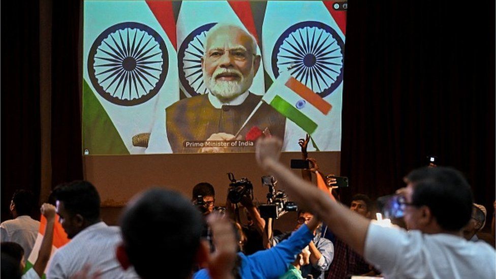 People wave India's national flag as India's Prime Minister Narendra Modi (C screen) congratulates the Indian Space Research Organisation (ISRO) for the successful lunar landing of Chandrayaan-3 spacecraft on the south pole of the Moon during a live stream of the event at the Nehru Science Centre in Mumbai on August 23, 2023.