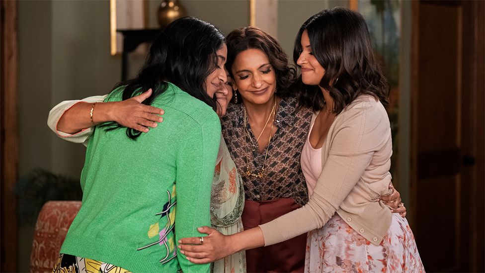 Characters of Kamala, Devi, Nalini and Nirmala hugging each other. Nalini is wearing a checked top and maroon trousers. Kamala is wearing a pink top, grey cardigan and floral skirt. Devi is wearing a light purple top, green cardigan and a patterned skirt. The three of them are covering Nirmala in a tight embrace.