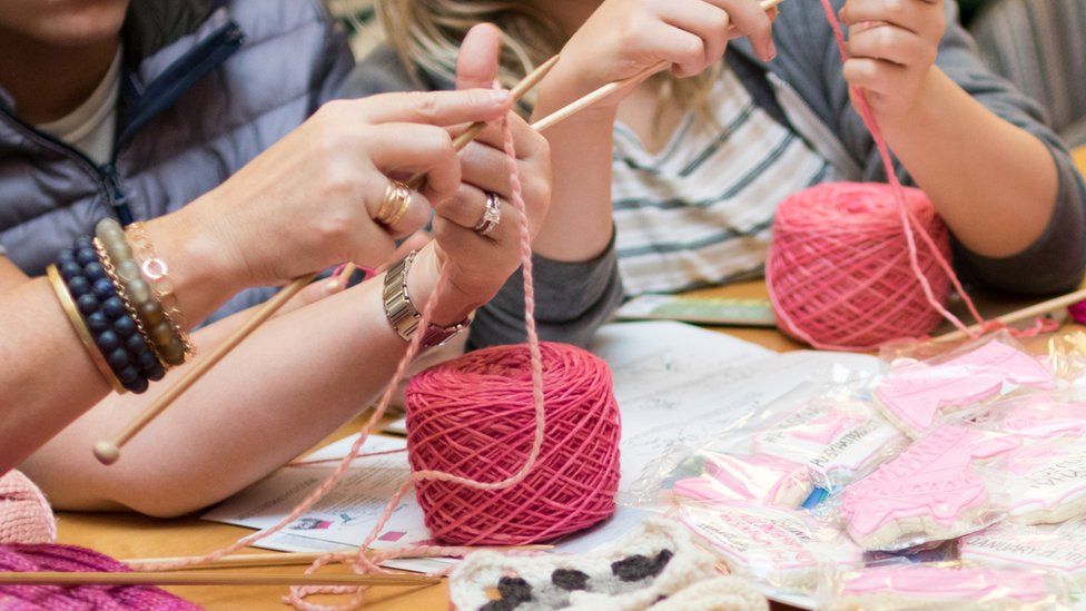 Women's hands can be seen starting a fresh knit from two balls of bright pink yarn