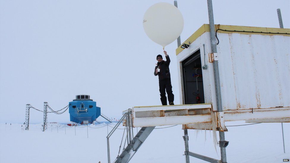 Peter Gibbs standing outside a small shed, holding a large white balloon in his hand. Halley research base lies in the background. Around is pure white snow and the sky is white
