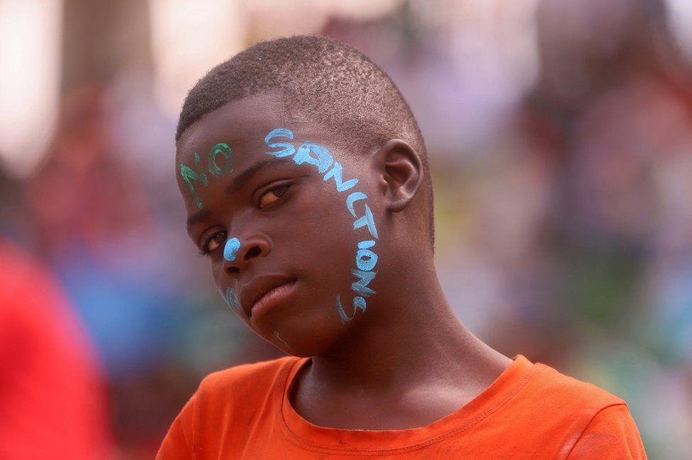 A boy with "no sanctions" daubed on his face in greena nd blue paint looks at the camera.