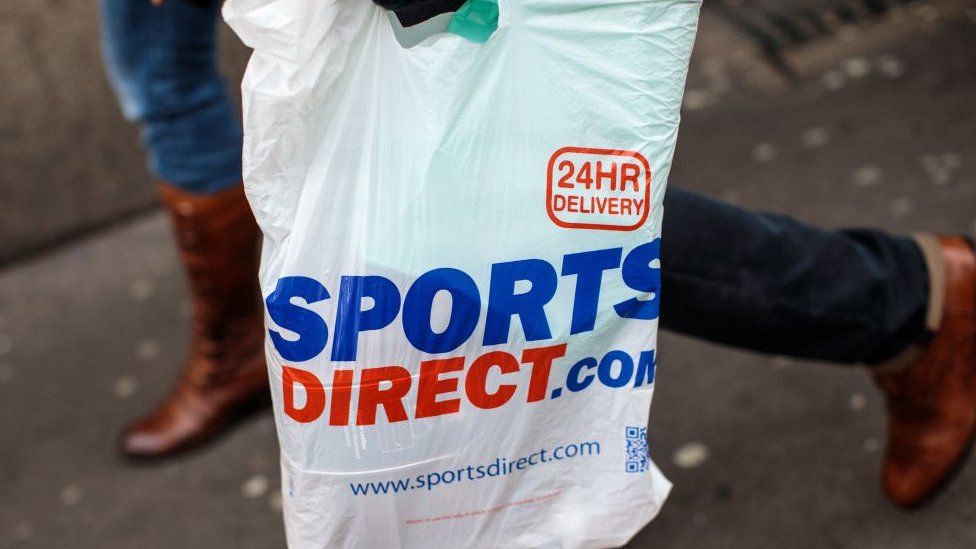 A plastic Sports Direct bag is carried by a shopper.