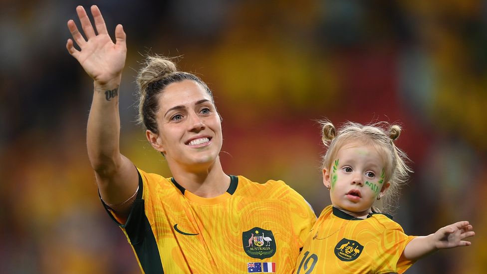 Katrina Gorry carries two-year-old daughter Harper, while wearing matching football kits, as they wave to the crowd